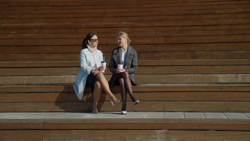 Two Attractive Woman Seating on the Bench, Talking and Drinking Coffee in the Center of the City
