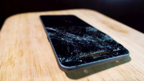 Smashed glass of mobile phone on wooden table rotating in light with half the screen broken and the frame bent