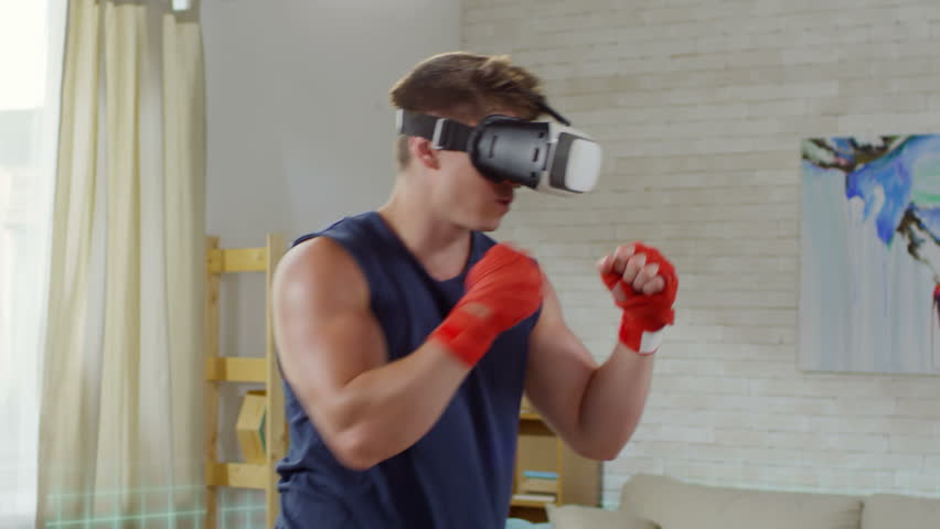 Young muscular man in VR headset with projected physical activity metrics boxing with holographic opponent while training in the living room Royalty-Free Stock Footage #1017531538