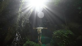 Young man standing arms outstretched in tropical rainforest, beautiful light coming from above making sunbeams- 4K video- people travel nature beauty concept 