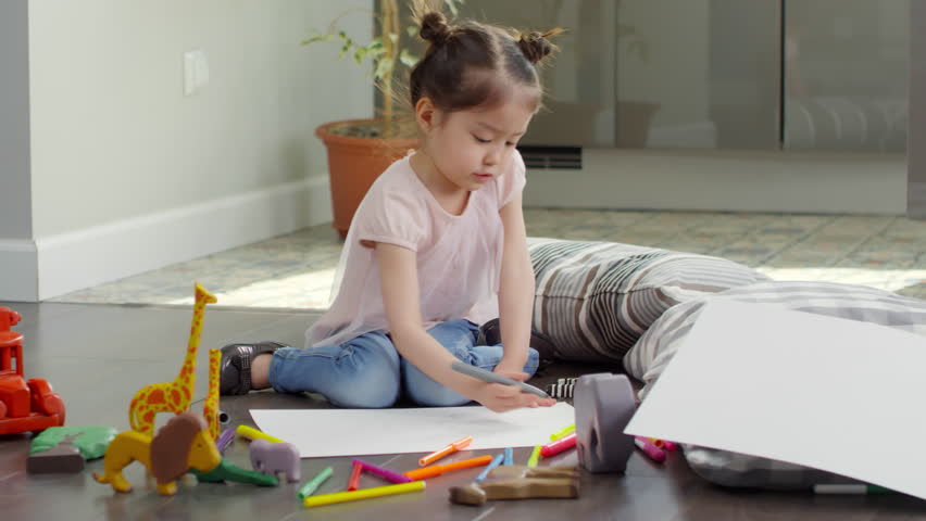 Tracking shot of cute Asian preschool-age girl sitting on floor and drawing on sheet of paper with color pencils