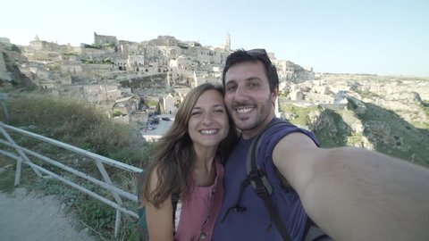 Beautiful tourist couple in Matera Italy taking selfies pictures with smartphone on a viewpoint with landscape. Travelling in Basilicata, famous site of stones of Matera, Italy