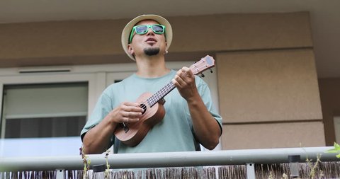 Coronavirus, COVID-19. Funny man in a hat and sunglasses plays the ukulele. Cheerful artist performs on the balcony in the summer.