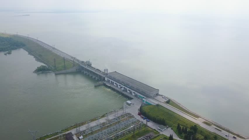 Novosibirsk Hydroelectric Power Plant is a hydroelectric power station on the Ob River in the Soviet district of the city of Novosibirsk. The concrete dam and powerhouse complex span the river, flanked by long earthen embankments that extend across the broad water surface. The structure features a low-profile concrete spillway section with multiple bays and an adjacent powerhouse building, all viewed from an elevated aerial perspective. The surrounding landscape is dominated by the expansive, calm reservoir on one side and the regulated downstream channel on the other, with an electrical substation visible in the foreground. The scene is captured under overcast skies with soft, diffused daylight, creating a muted palette of greens, greys, and pale blues. The camera maintains a steady, sweeping aerial orbit that reveals the full scale of the installation and its integration into the flat West Siberian terrain. The facility is the only large-scale hydroelectric plant on the Ob River, commissioned in 1957 and visited by both Nikita Khrushchev and Richard Nixon in 1959.