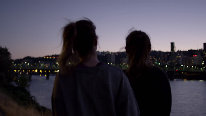Friends Walk Along River At Night, Woman Holds Up A Peace Sign Briefly, City In Background