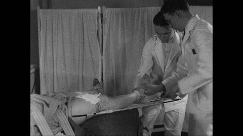 1930s: UNITED STATES: doctor bandages patient's leg in splint. Patient with leg in splint