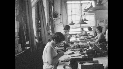 1920s: Women workers sit and stand at tables, folding and boxing stockings. Stacks of boxes. Women fold manufactured clothing in mill.