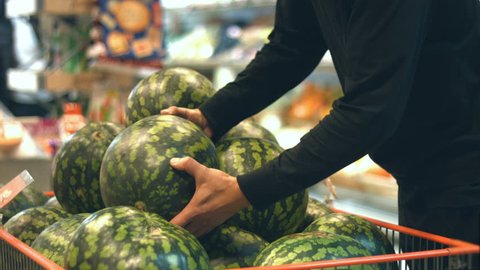 Fruit box with watermelons at supermarket.
Man moves with basket to box with watermelons then men's hands take watermelon and put it in the basket.