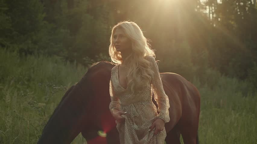 Portrait of a beautiful blonde model in open field with horse in summer, sunset light. Girl in dress posing outdoor, brown horse on a background.