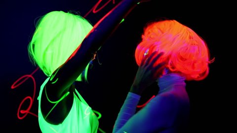 Beautiful sexy women with laser, UV face paint, wig, glowing glasses, glowing clothing dancing together fast in front of camera, Half body shot. Caucasian and asian woman. Party concept.