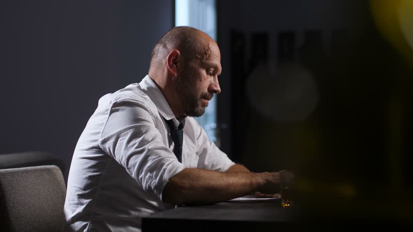 Emotional stressed middle aged businessman reviewing financial documents at desk in office. Distraught, overloaded with company debts entrepreneur loosing his necktie and looking hopelessly indoors.