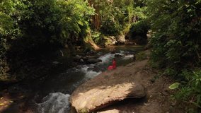 4K aerial drone video of young woman in red dress sitting on the rocks close to the mountain river. Bali island.