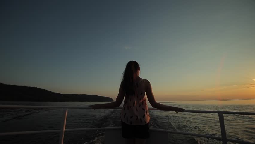 A beautiful girl stands in the wind against sunset sky, river, and mountains. Back view