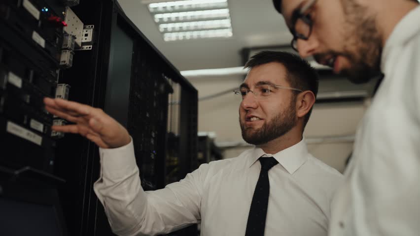 Two IT engineers are working in a data center with rows of server racks and super computers. They are discussing their work as they check cables and other equipment. Royalty-Free Stock Footage #1017667870