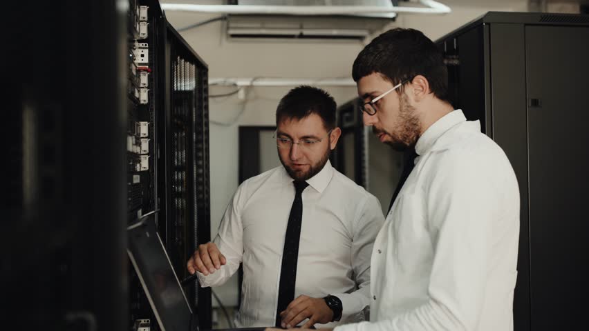 Two IT engineers are working in a data center with rows of server racks and super computers. They are discussing their work as they check cables and other equipment. Royalty-Free Stock Footage #1017667876