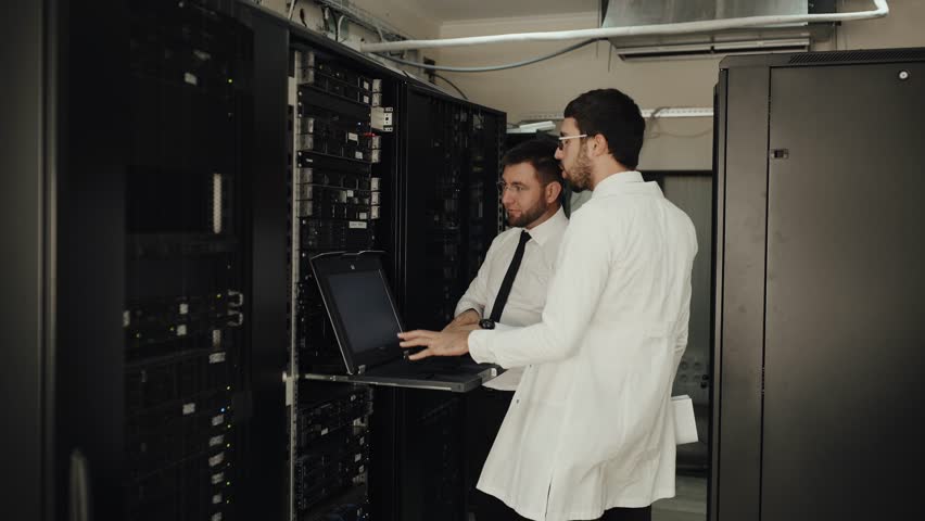 Two IT engineers are working in a data center with rows of server racks and super computers. They are discussing their work as they check cables and other equipment. Royalty-Free Stock Footage #1017667885