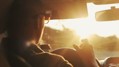 A close back view of a grown-up man in sunglasses driving a car during daytime. Bright sun shines in his eyes