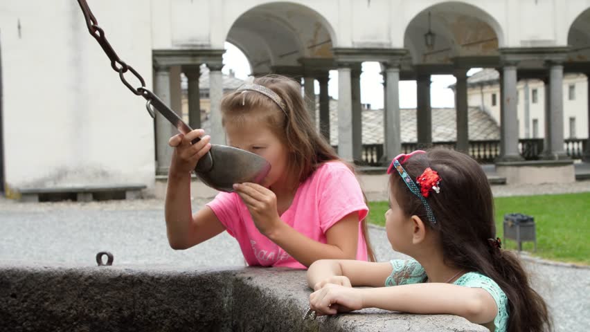 OROPA, BIELLA, ITALY - JULY 7, 2018: girls kids drink silver alpine water from silver buckets, from stone gothic mask fountain in Shrine of Oropa, Sanctuary, in the mountains near the city of Biella