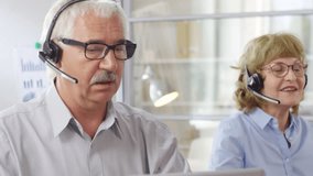 Senior male and female support managers in headsets talking with clients via video call and typing on computers while working together in call center
