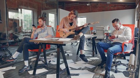 Business group of young people are bored in the office without work after a failure of business meeting. Woman with guitar setting on table, two mens playing with a phones, paper is scattered in floor