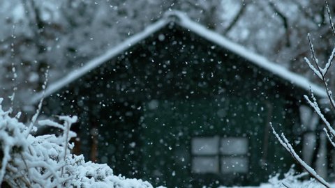 SLOW MOTION, DOF: Cinematic shot of a wooden shack in the woods on a beautiful snowy day. Lonely green hut in the middle of the forest weathering the snowfall during a cold winter. Winter wonderland.