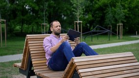 Young man watching movie on tablet sitting on bench in city park

