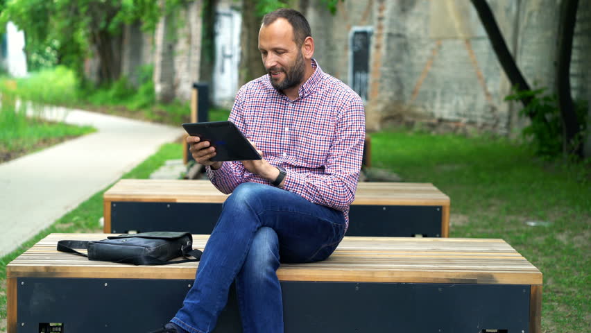 Young man watching movie on tablet sitting on bench in city park
