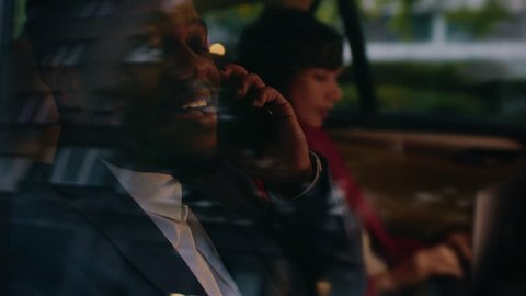 Beautiful Businesswoman and Handsome Businessman Riding on the Backseat of a Car in the Evening. Man Makes a Phone Call, Woman Works on a Laptop. Camera Shot made from Outside the Car.