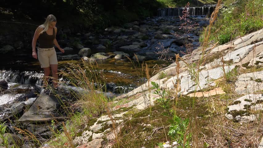 A young beautiful woman climbs up a slope near a creek in a forest on a sunny day