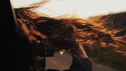 Close view of girl s long hair waving by wind and her hands playing with them while she is riding a car and looking out of the window. Rear view