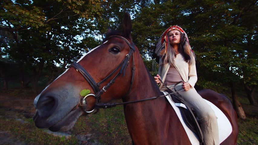 Pretty independent woman in native american indian headdress is riding brown horse in the forest. Sunny autumn day