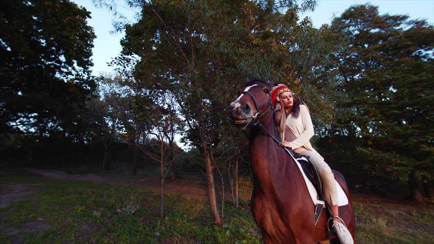 Sliding veiw of young strong woman in native american indian headdress riding brown horse. Sunset. Green trees are on the background