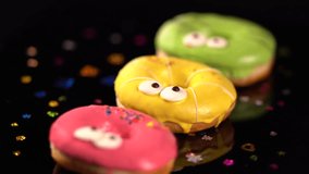 Three spinning colorful iced ring donuts with fun googly eyes and scattered confetti on a dark background