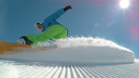 SLOW MOTION, LOW ANGLE, LENS FLARE: Cheerful young male snowboarder carving a freshly groomed slope in the winter sun. Cool shot of snowboarder doing a cool hand drag while riding in the sunny Alps.