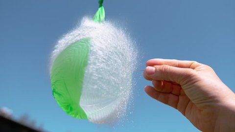 SLOW MOTION, CLOSE UP: Unrecognizable person holding a needle pierces a green water balloon. Hand holding a metal pin bursts a rubber balloon filled with a crystal clear liquid. Water droplets flying.