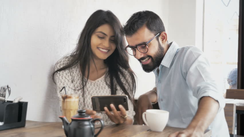 A young couple are laughing while watching a funny video on a smartphone or cellphone sitting in modern coffee shop. Two happy and joyous friends watching a movie on a mobile phone in a cafe interior  Royalty-Free Stock Footage #1017802600