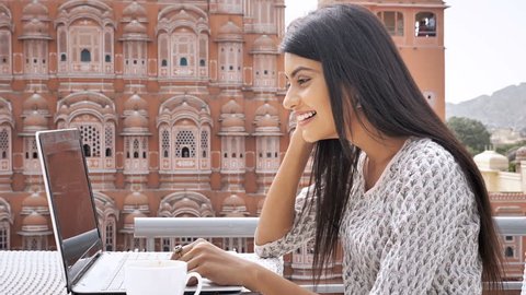 A happy and cheerful young woman in traditional kurta is using laptop to talk on video call. A  movement shot of a smiling and attractive girl chatting on video call with a friend in exterior setup 
