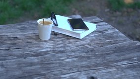 Book, glasses, coffee and mobile on an old wooden table outdoors in a park in a revolving view