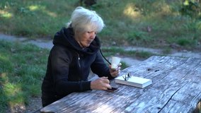 Woman sitting at an old wooden table drinking coffee in a park using her mobile phone with a book and reading glasses alongside