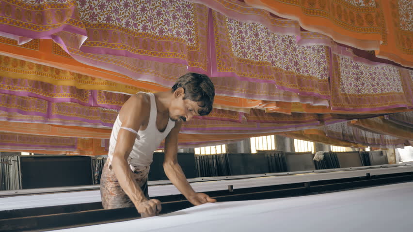 A movement shot of a daily wage/ migrant worker/ laborer/ migrants working in textile printing interior factory setup while colorful printed cotton sheets hanged on the top to let them dry Royalty-Free Stock Footage #1017820057