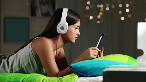 Side view portrait of a teen e-learning with a tablet and headphones lying on a bed in the night at home