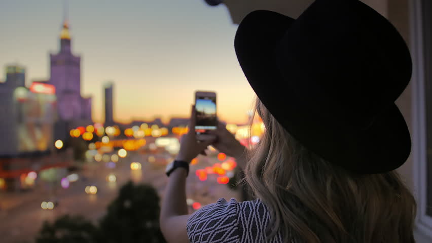 The girl in the hat makes a photo of the night city. Uses a smartphone. In the frame of a skyscraper in the center of the capital. Warsaw.