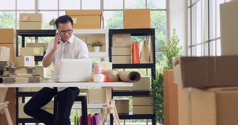 Young Asian businessman in casual shirt look at notebook among several parcels,  working in simple house office look like doing startup business. Concept for online marketing, SME and home workplace  Vídeo Stock
