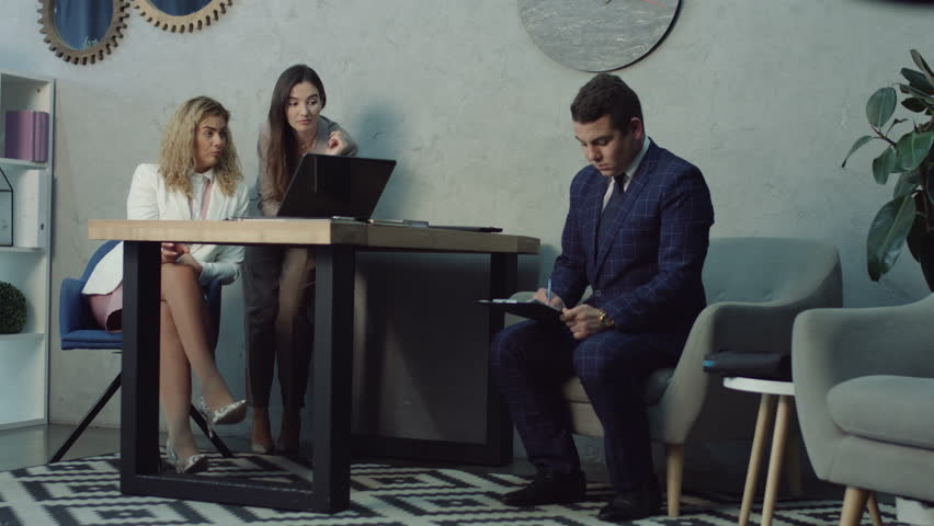 Rude gossip female colleagues humiliating and offending upset young male coworker at workplace. Two giggling businesswomen bullying sad colleague and whispering about man in office. | Shutterstock HD Video #1017840538
