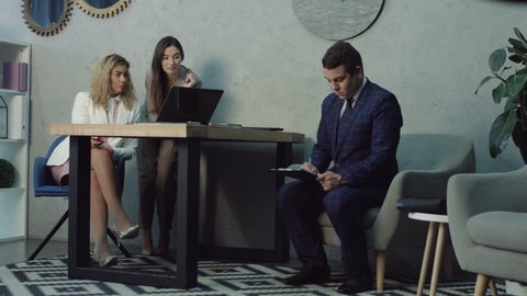 Rude gossip female colleagues humiliating and offending upset young male coworker at workplace. Two giggling businesswomen bullying sad colleague and whispering about man in office.