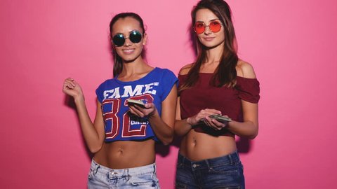 Two young sexy stylish smiling brunette women making it rain money. Hot girls dressed in summer hipster clothes and throwing bills out of a bundle money. Posing on pink background. 4k