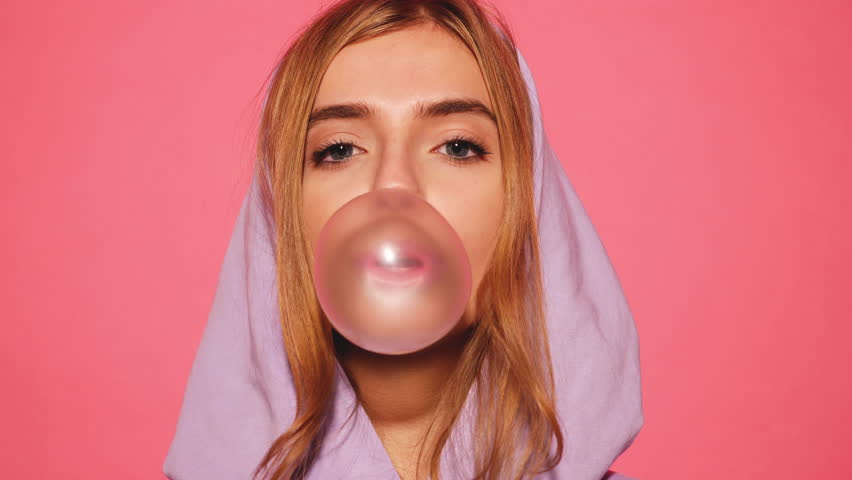 Portrait of young sexy playful blond girl dressed in blue hoodie standing and isolated over pink background blowing bubblegum candy bubbles and chewing gum. Woman looking at camera. 4k. Slow motion