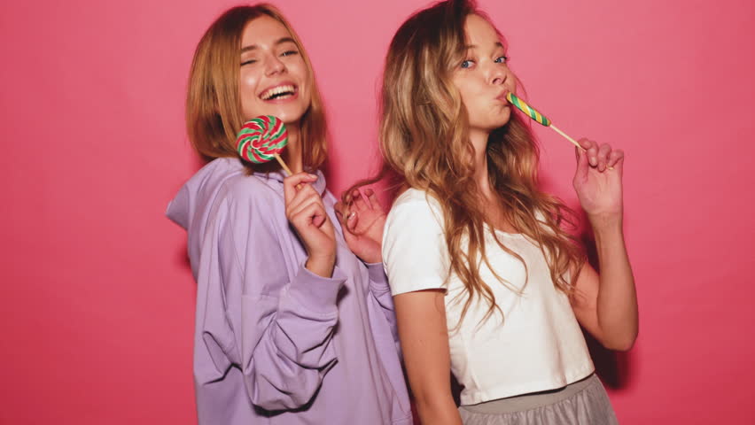Two young beautiful smiling blond hipster girls in trendy summer clothes. Carefree hot women posing near pink wall. Positive funny models hugging and eating lollipop.  4k Royalty-Free Stock Footage #1017854251