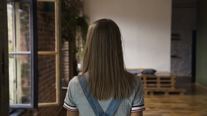 Cute hipster girl with earphones around the neck is walking with great pleasure after successful ride with skateboard looking at the camera and smiling, slow motion