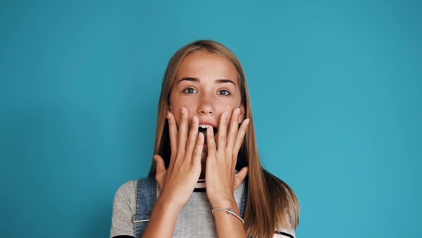 Happy girl with long hair laughing and keeping hands at her face while standing in indoors of isolated over blue background. Portrait of beautiful surprised and shocked teen girl Emotion, lifestyle